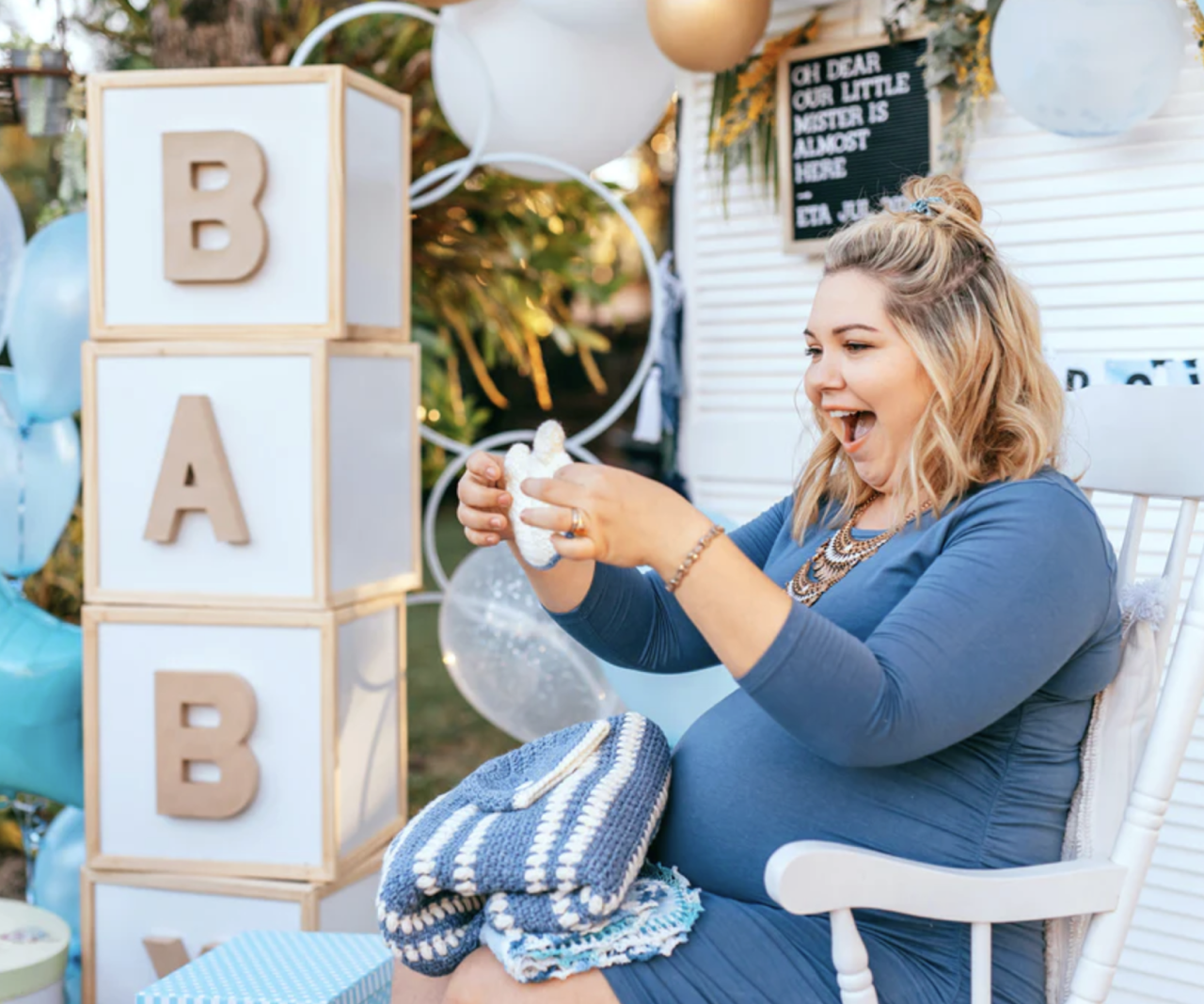 A pregnant woman in a chair holding up knit baby clothes.