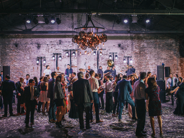 Large group of people dancing and talking with each other in a warehouse while a band plays.